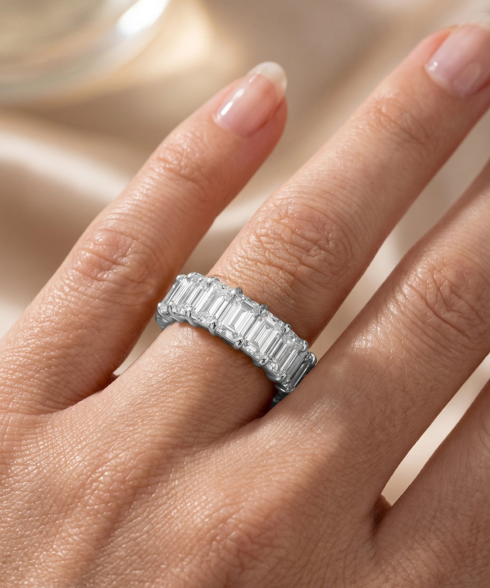 A close-up of a hand wearing a silver band ring with a row of baguette-cut clear stones, set against a soft beige satin background.