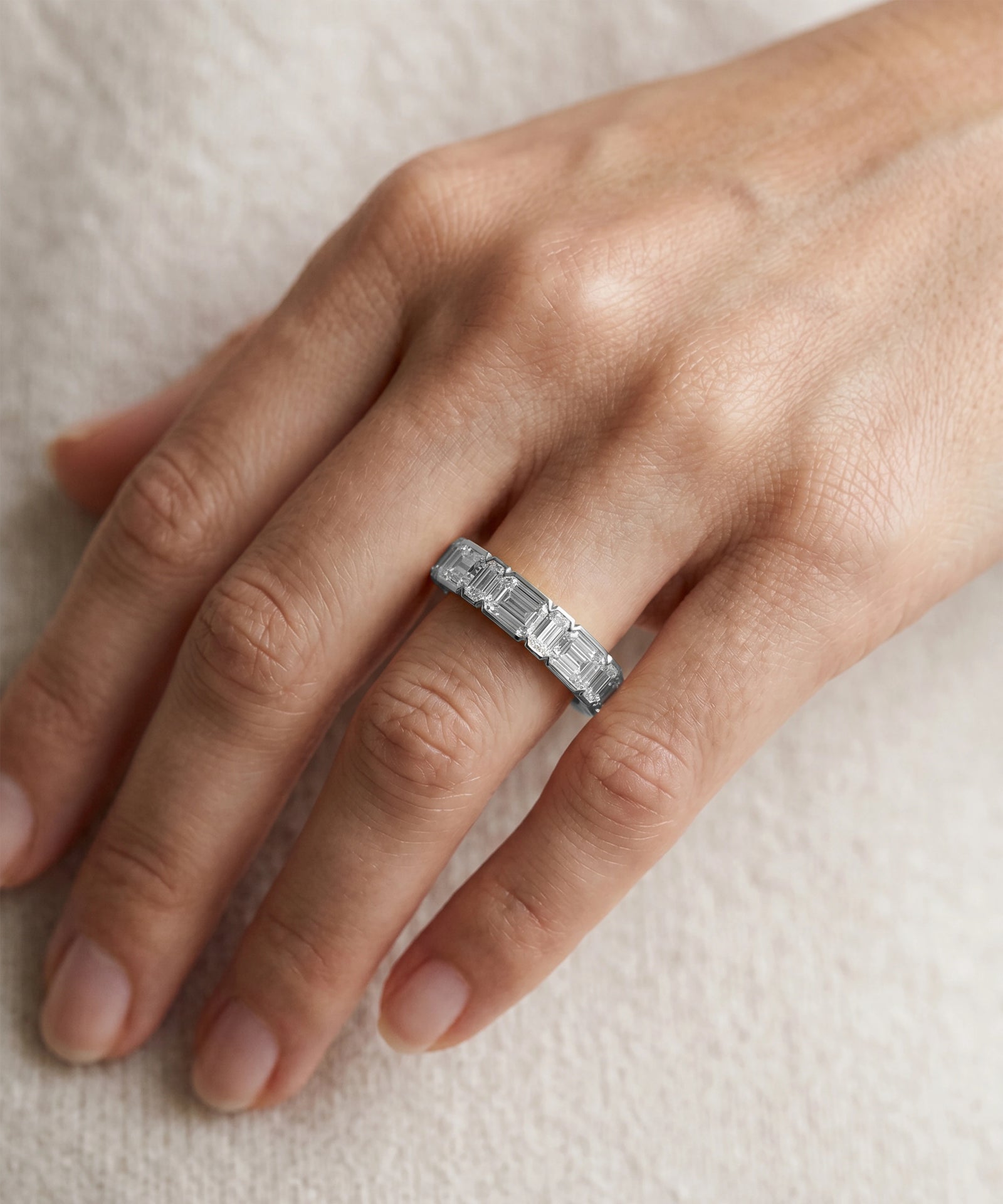A close-up of a hand wearing a silver band ring with baguette-cut clear stones, resting on a soft beige fabric background.