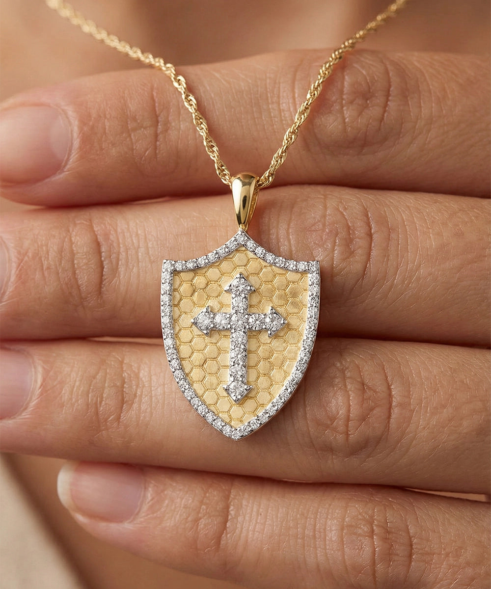 Gold necklace with a shield-shaped pendant featuring a cross design, held between fingers.
