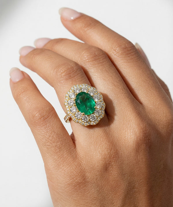 Hand wearing a ring with an emerald and diamond halo on a light background