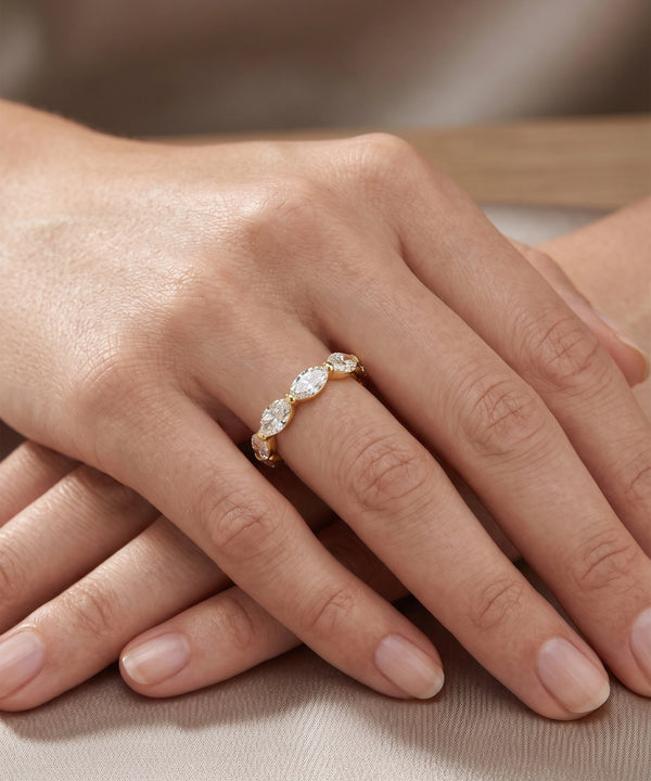 A close-up of a hand wearing a delicate gold ring with three marquise-cut clear gemstones, resting on a neutral beige fabric surface.