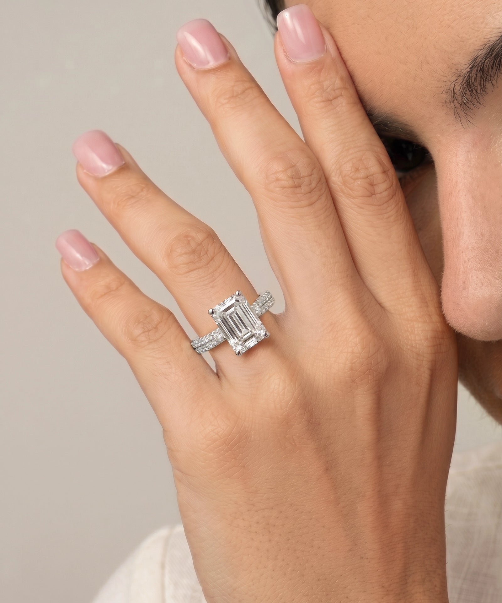A close-up of a person's hand wearing a silver ring with an emerald-cut clear center stone and small side stones, against a soft light gray background.
