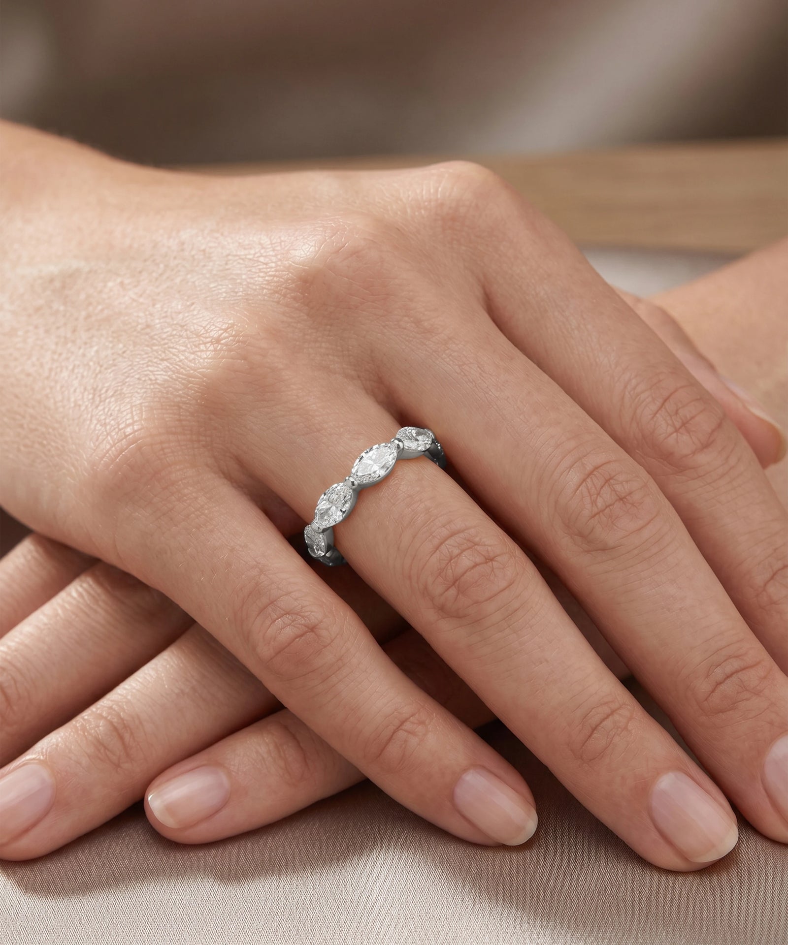 A close-up of a hand wearing a silver ring with marquise-cut clear gemstones, resting on a neutral beige fabric surface.