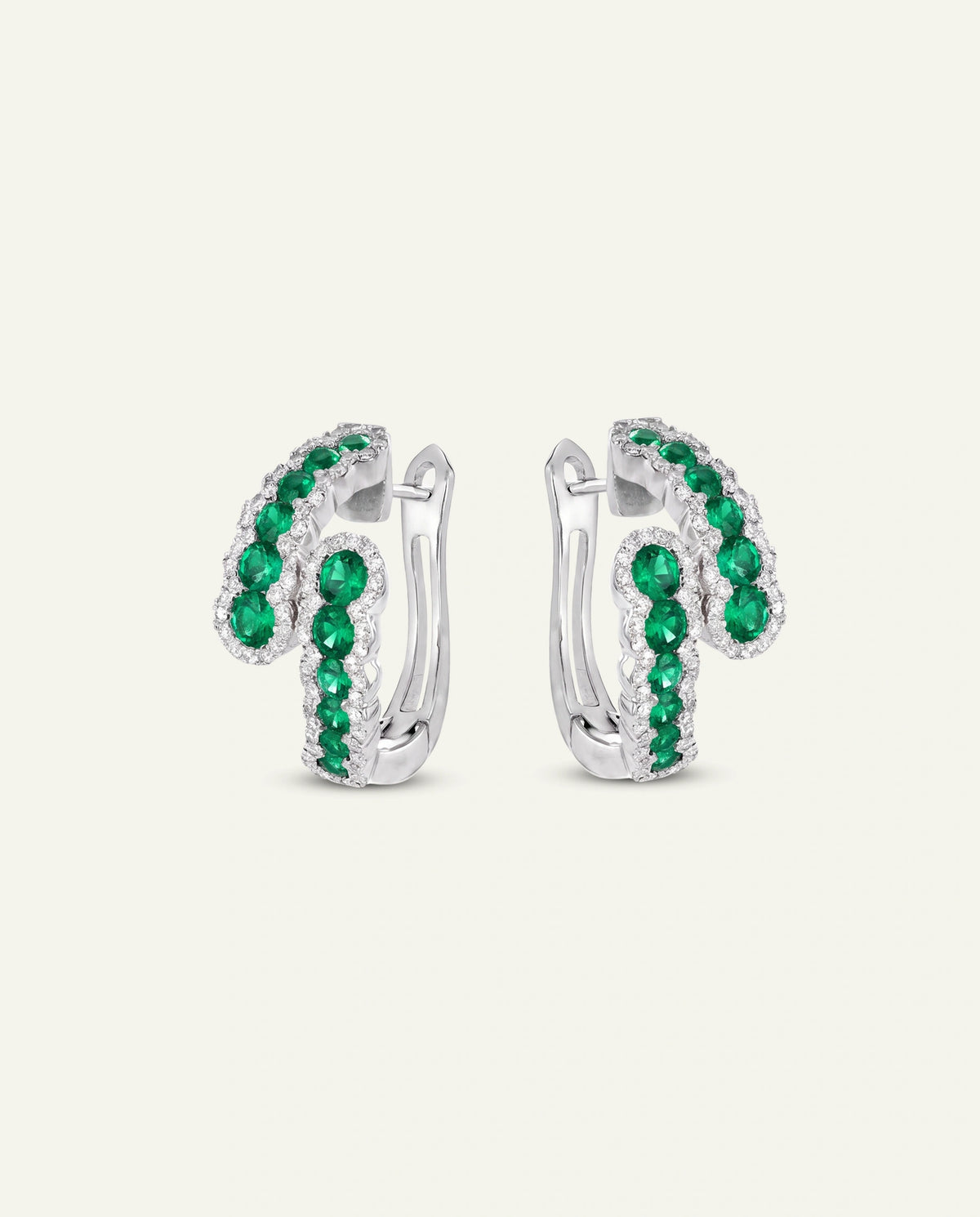 A pair of silver huggie hoop earrings adorned with green emerald-cut gemstones and clear round stones, set against a solid off-white background.