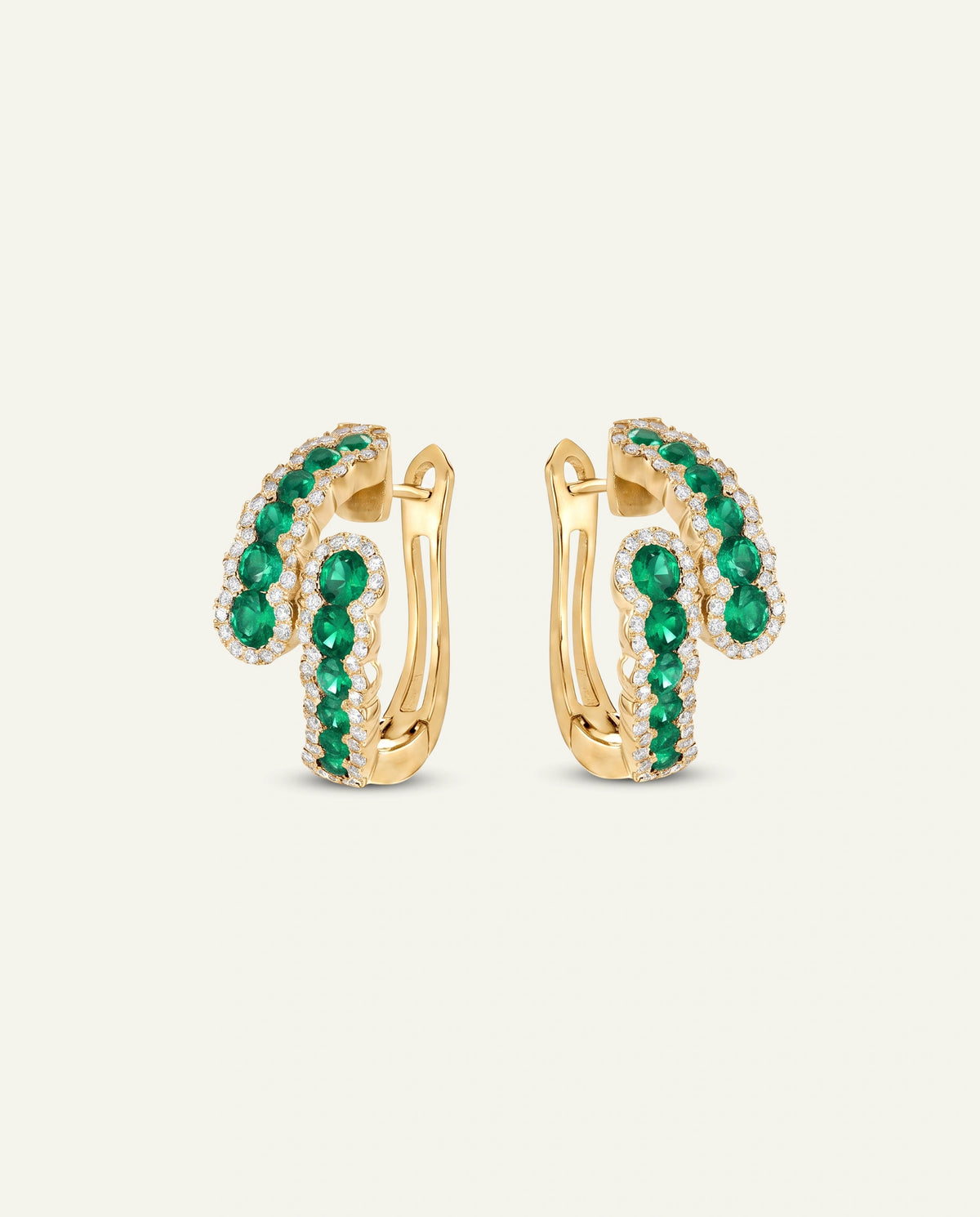 A pair of gold huggie hoop earrings adorned with curved rows of round-cut green emeralds and small clear stones, shown on a light cream background.