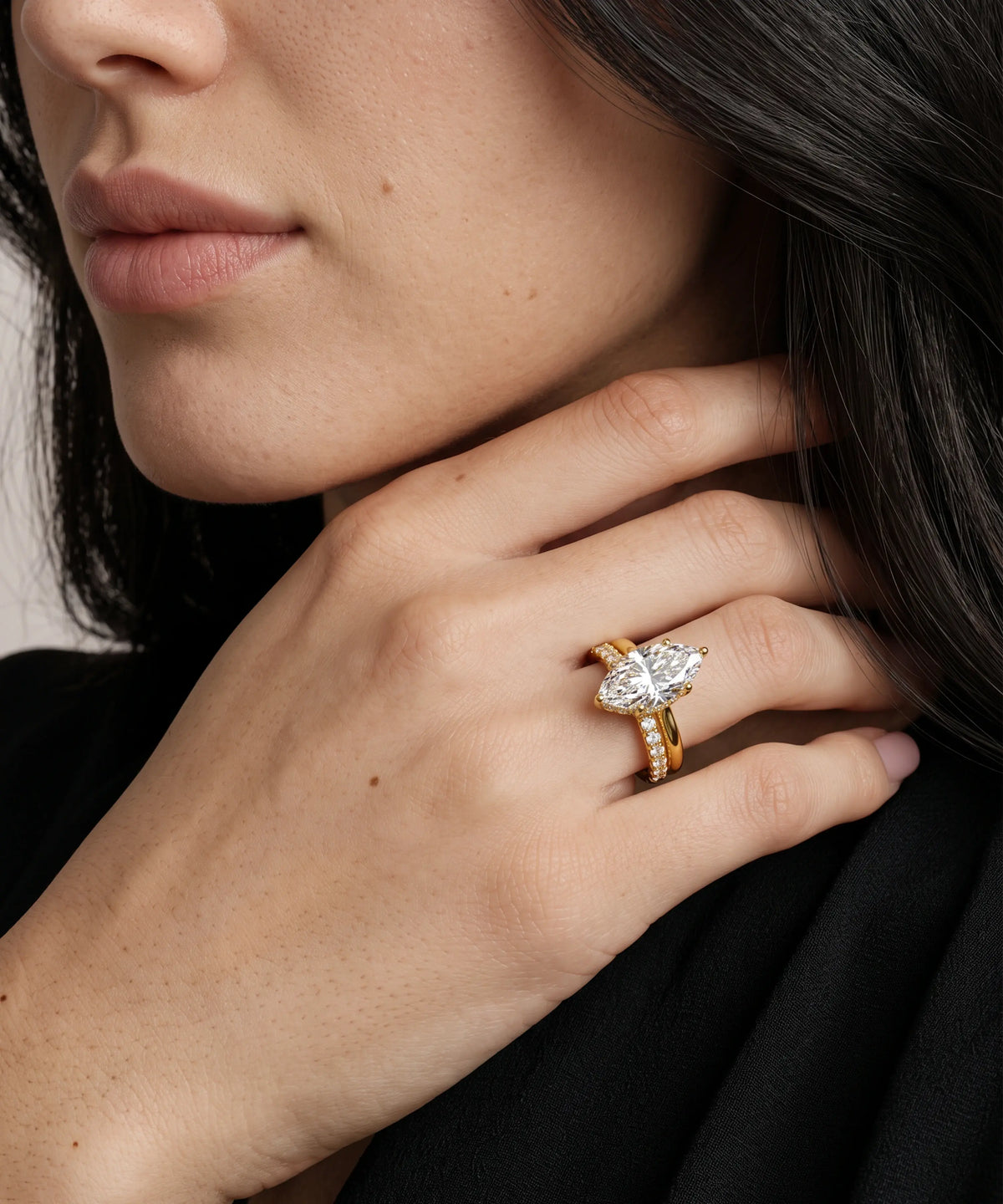 A close-up of a model's hand wearing a gold ring with a marquise-cut diamond and a smaller gold band with pavé-set stones, against a dark background.