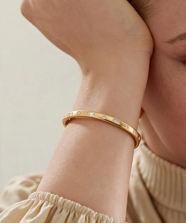 A close-up of a person's wrist wearing a slim gold bangle bracelet with small clear rectangular stones set along the band, against a soft light beige background.