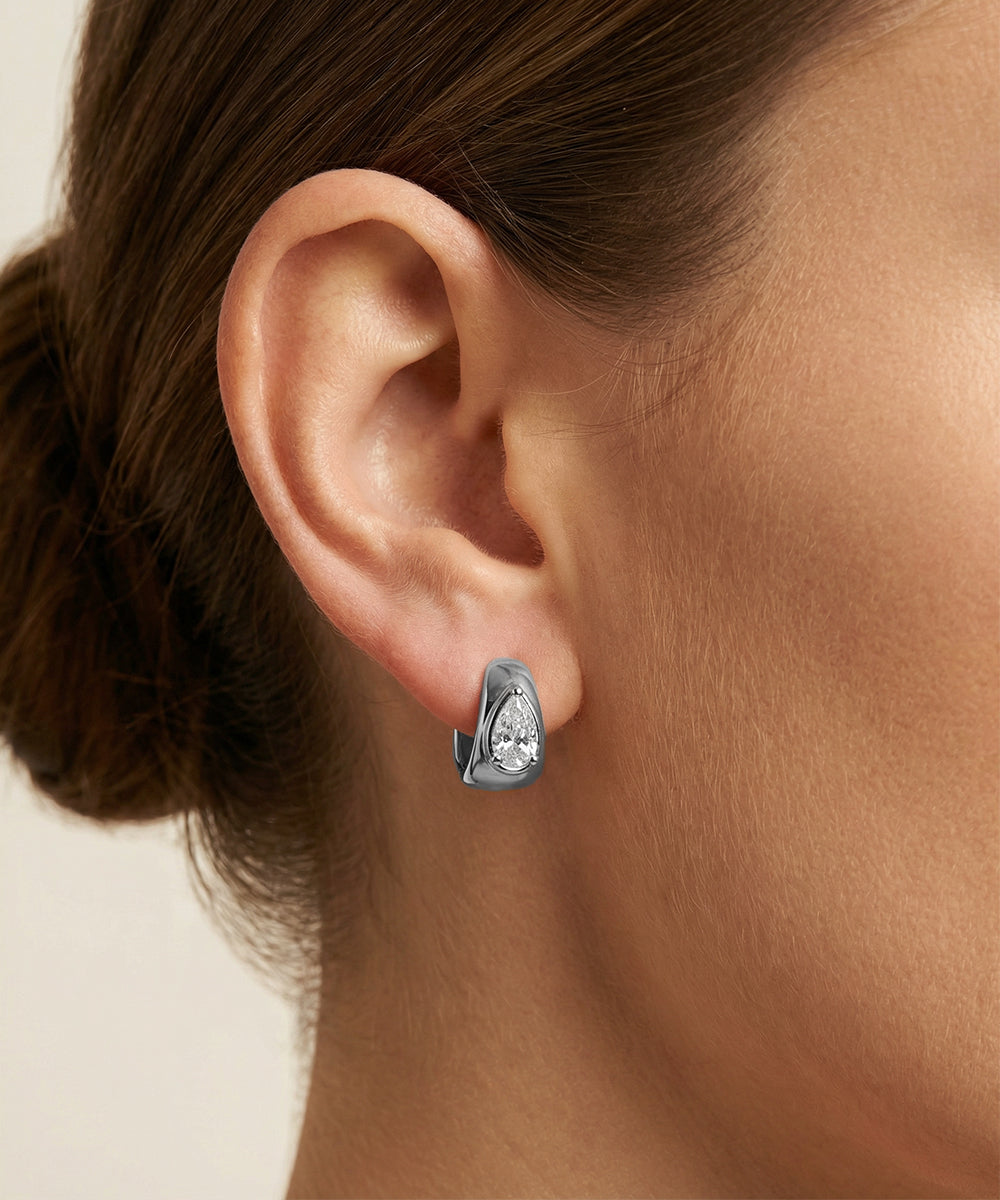 Close-up of an ear wearing a silver earring with a clear gemstone on a neutral background