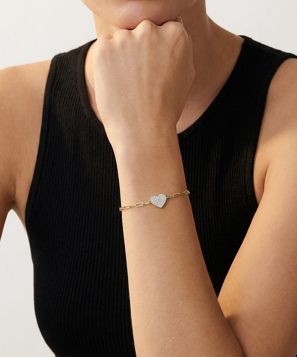 Person wearing a gold bracelet with a heart charm on a plain background
