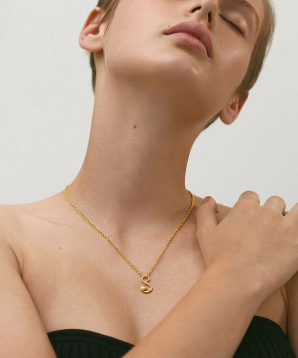 Woman wearing a gold necklace with a 'S' pendant on a plain background