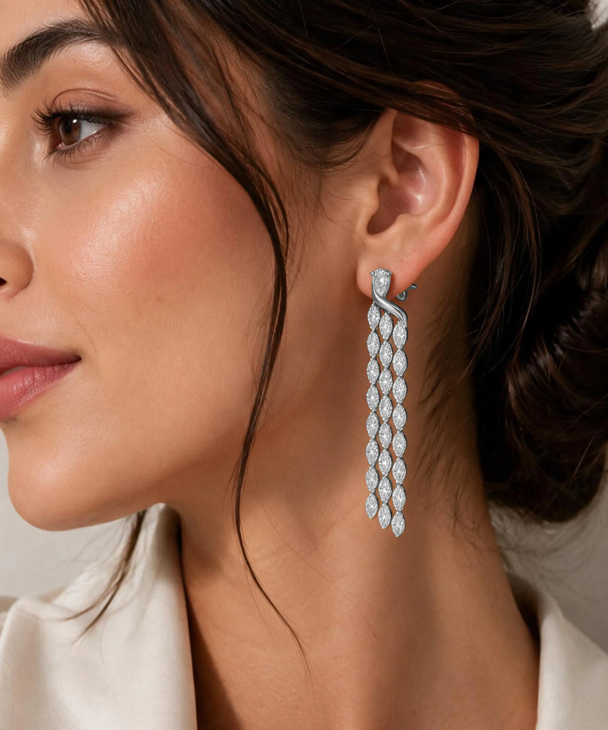 A close-up of a woman wearing a long, multi-strand dangle earring made of sparkling clear stones in a silver-toned setting. The earring hangs from a matching small stud earring with a tiny blue accent stone.