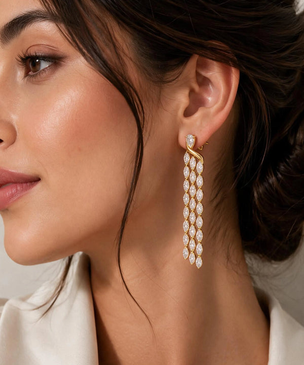 A close-up of a woman wearing a long, multi-strand gold dangle earring adorned with clear pavé gemstones. The earring hangs from a gold post and features five cascading strands of marquise-cut and round stones.