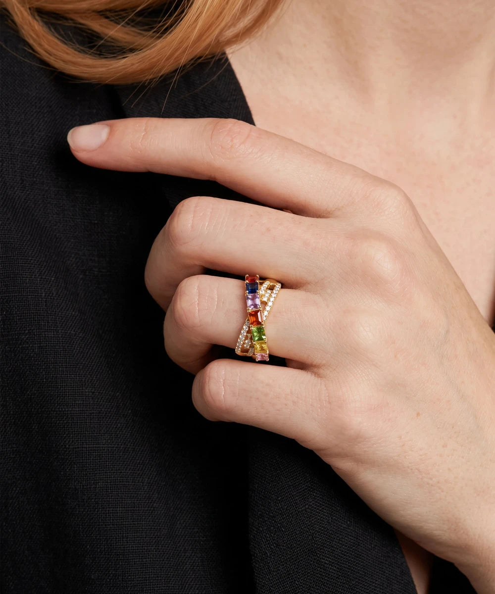 A close-up of a hand wearing a gold ring with multiple colorful gemstones, including blue, red, green, and orange, against a dark black top background.