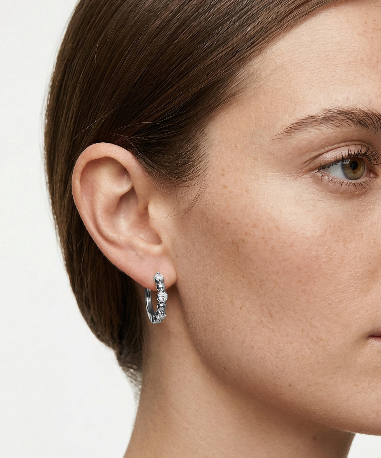 Close-up of a person wearing a silver hoop earring with small stones on a plain background