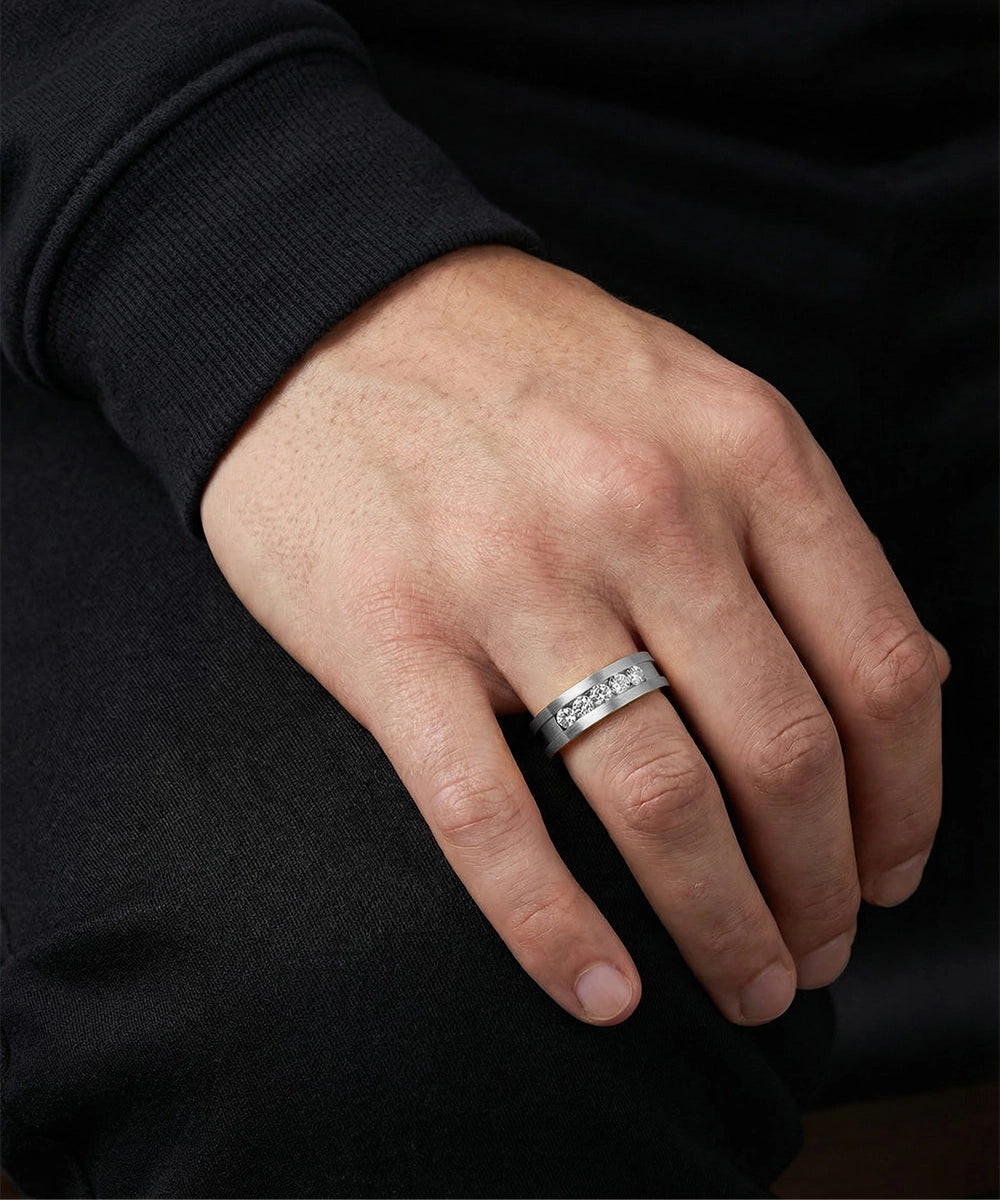 A close-up lifestyle shot of a man's hand wearing a wide platinum or white gold wedding band with a brushed matte finish and a central channel of inset round diamonds.