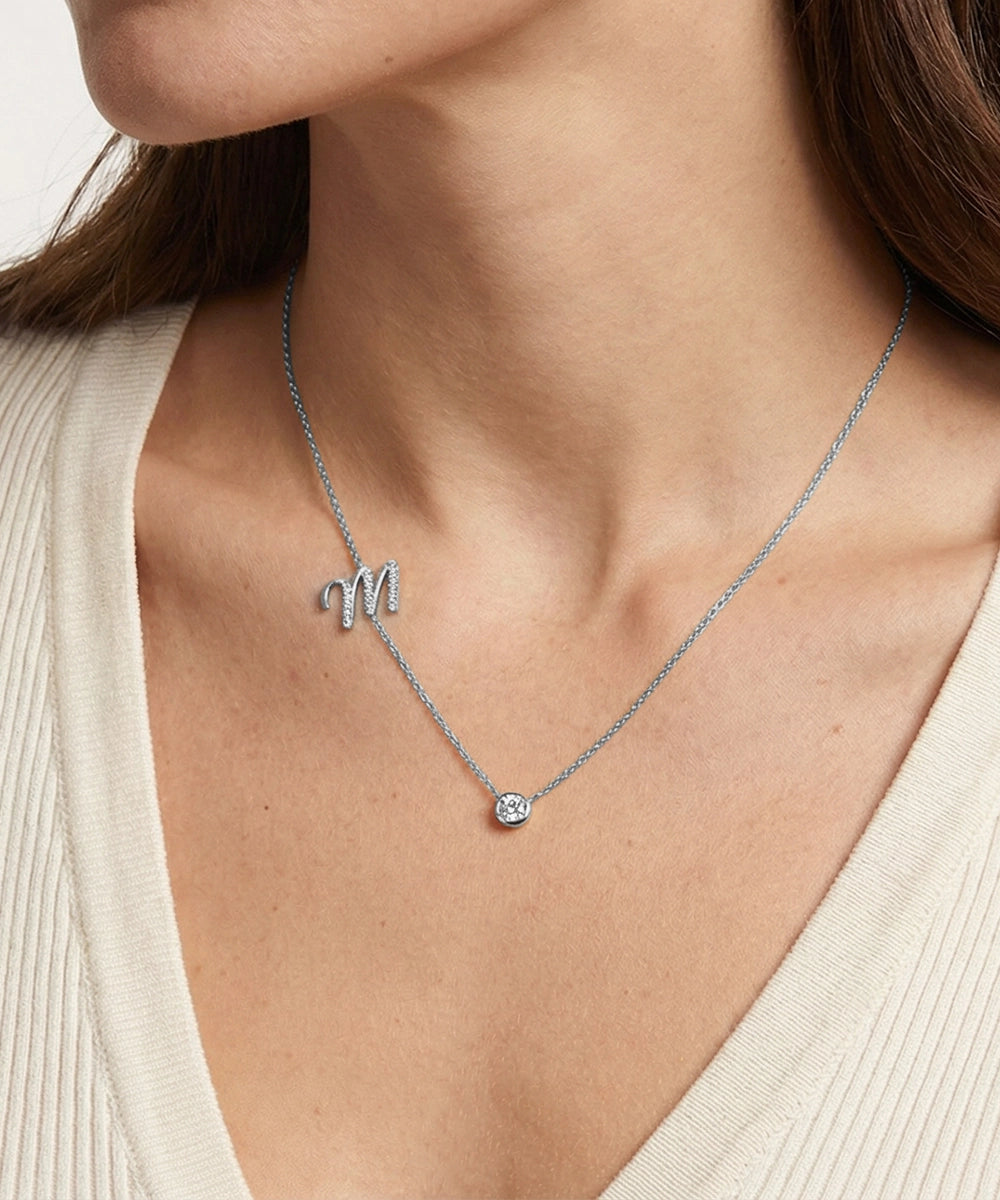 Necklace with a letter 'M' pendant worn by a person in a beige top.