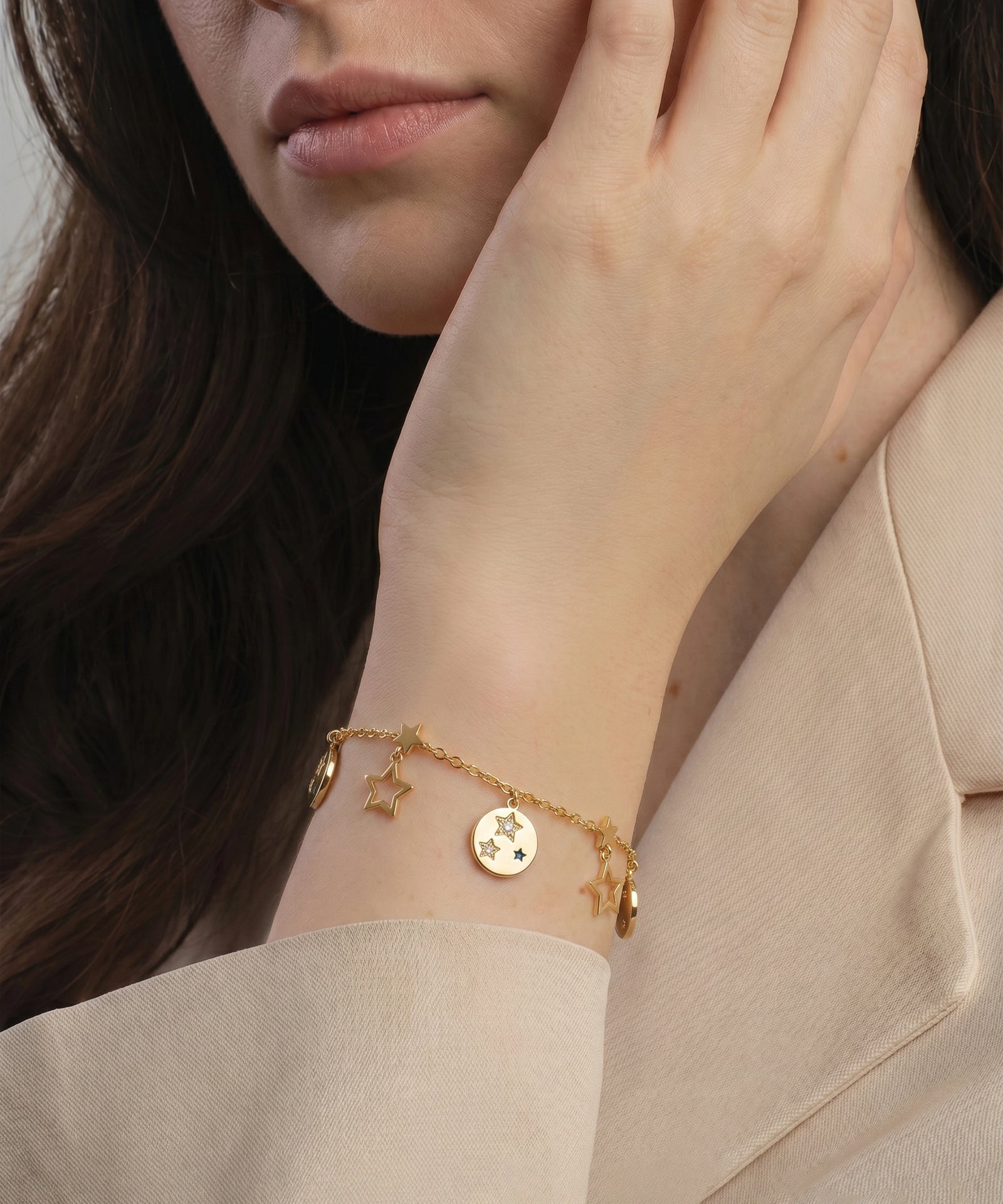 Close-up of a person wearing a gold bracelet with charms, set against a neutral background.