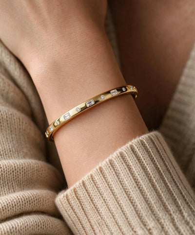 A gold bangle bracelet with rectangular clear stones and small black accents, worn on a person's wrist over a beige knit sweater. - alternate view