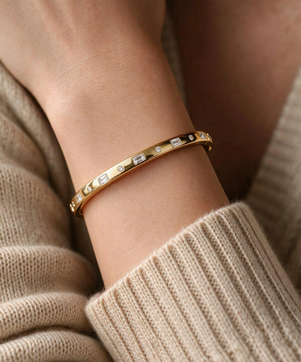 A gold bangle bracelet with rectangular clear stones and small black accents, worn on a person's wrist over a beige knit sweater.