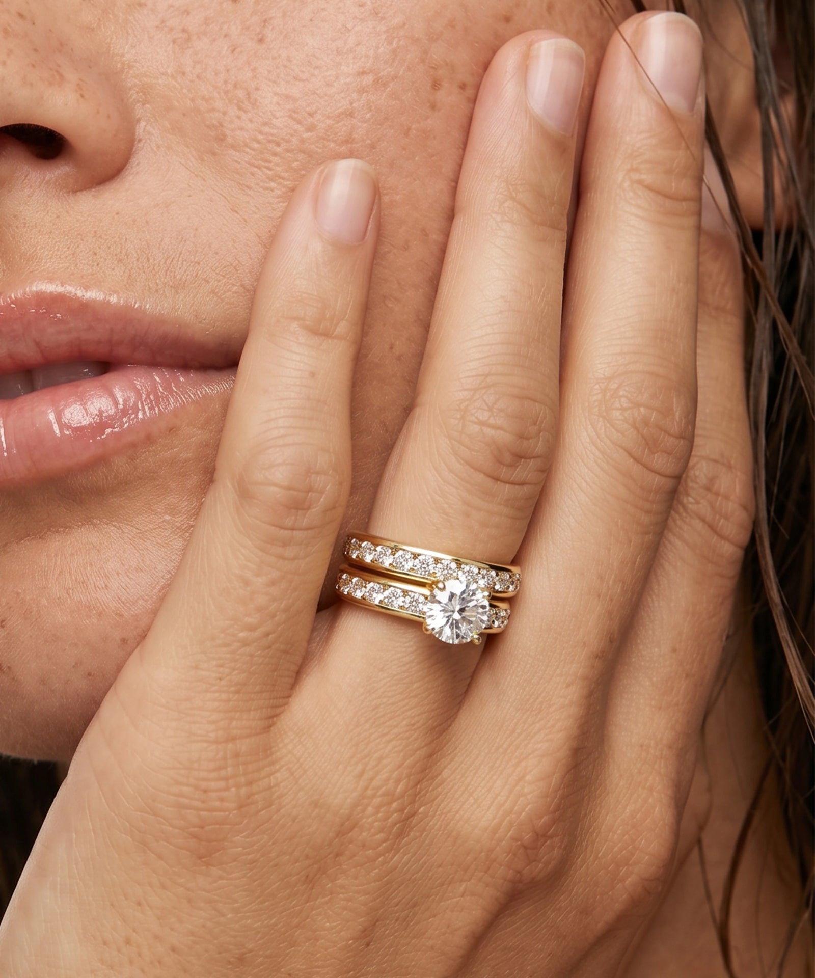 A close-up lifestyle shot of a model's hand resting against her cheek, showcasing a two-piece yellow gold diamond engagement ring set. The set includes a main ring with a round brilliant-cut center stone and matching pavé band, alongside a thinner band ring with a small round diamond.