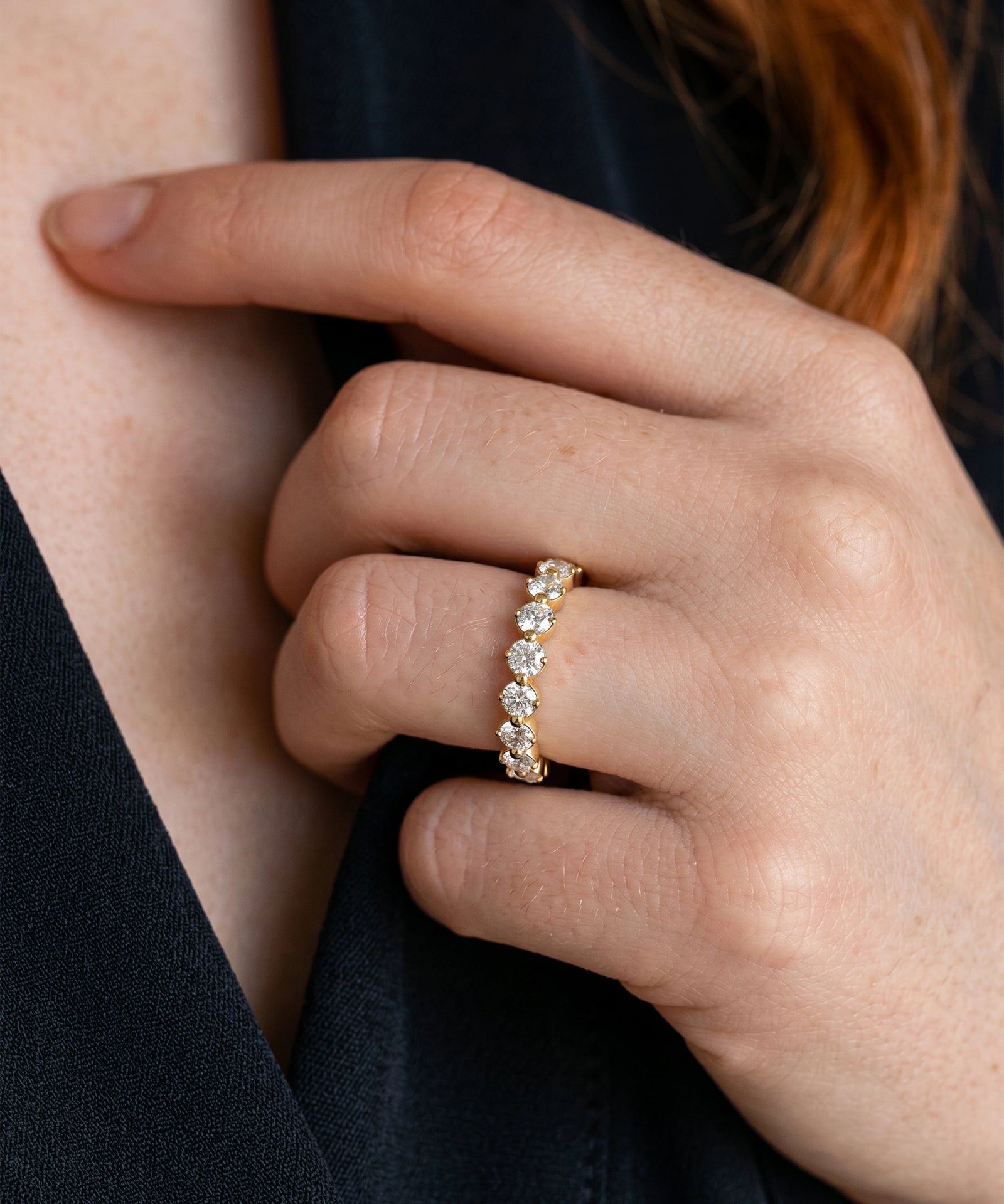 A close-up of a person's hand wearing a gold eternity ring set with clear round-cut gemstones, against a dark navy top and skin background.