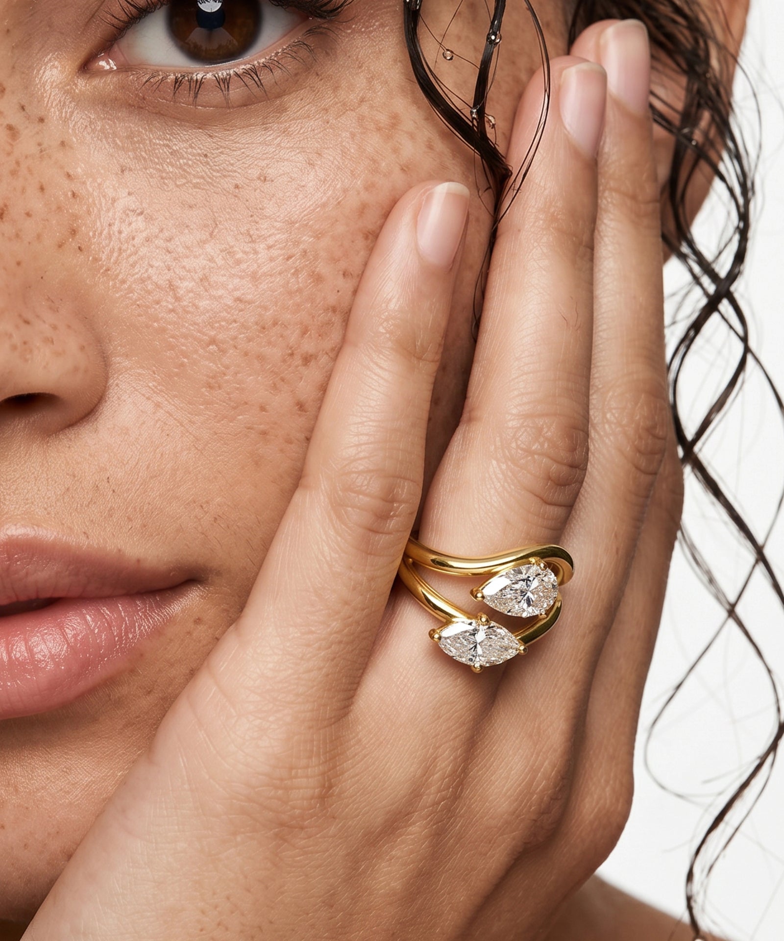 Close-up of a woman's hand with a gold ring featuring two pear-shaped diamonds, held near her face.