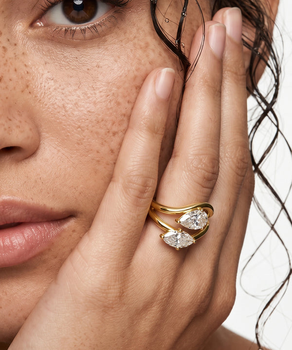 Close-up of a woman's hand with a gold ring featuring two pear-shaped diamonds, held near her face.