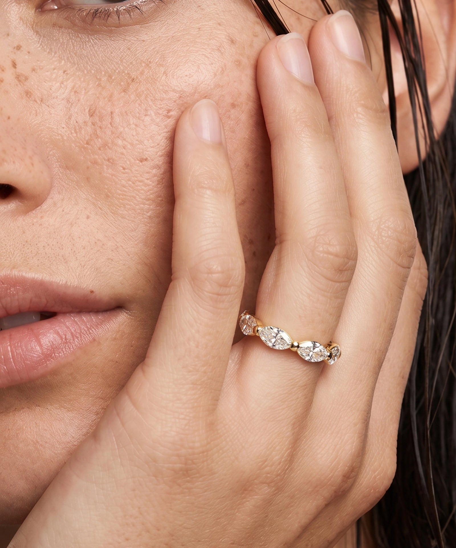 A close-up lifestyle shot of a model's face and hand wearing a delicate gold ring with marquise-cut clear gemstones.