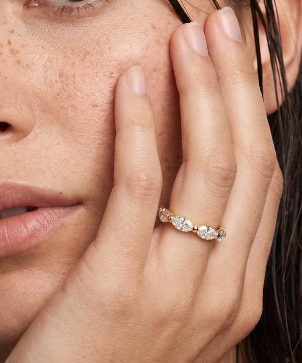 A close-up lifestyle shot of a model's face and hand wearing a delicate gold ring with marquise-cut clear gemstones.