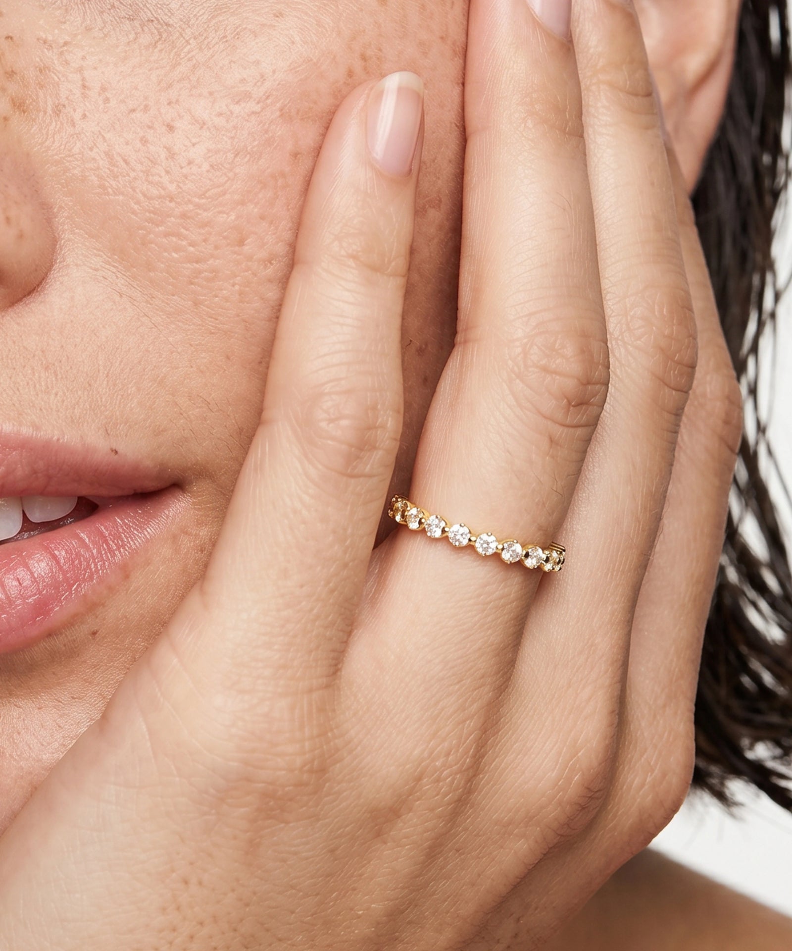 A close-up lifestyle shot of a woman's hand resting against her cheek, wearing a delicate yellow gold eternity ring set with small clear round-cut gemstones.