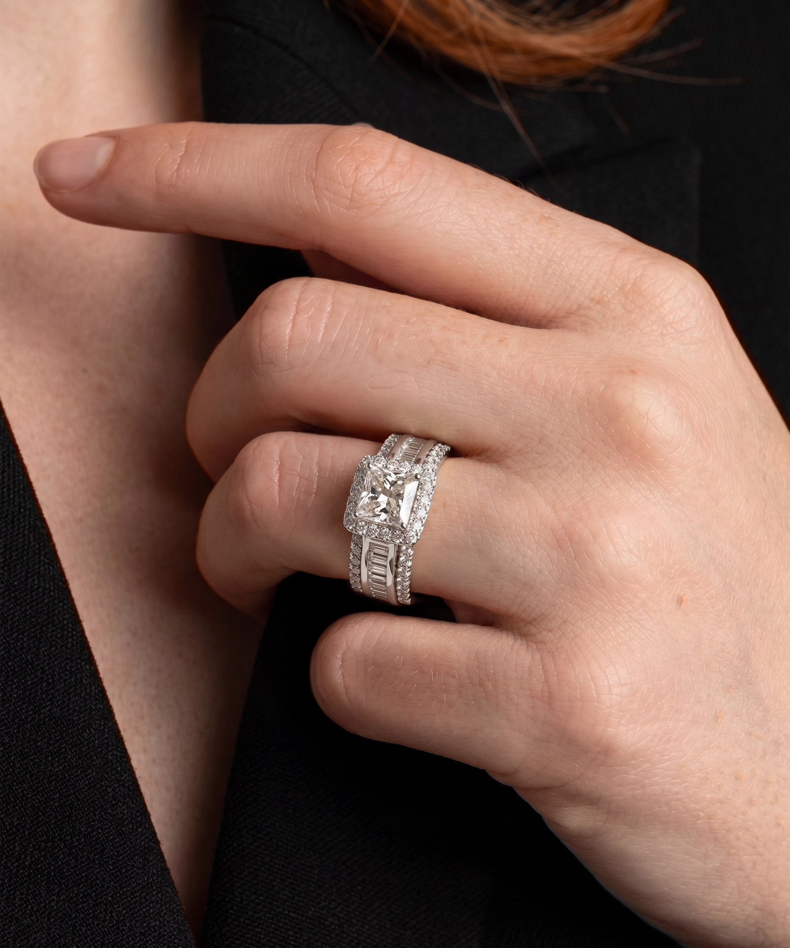 A close-up of a hand wearing a silver engagement ring with a round center stone and a matching band, set against a dark black blazer.