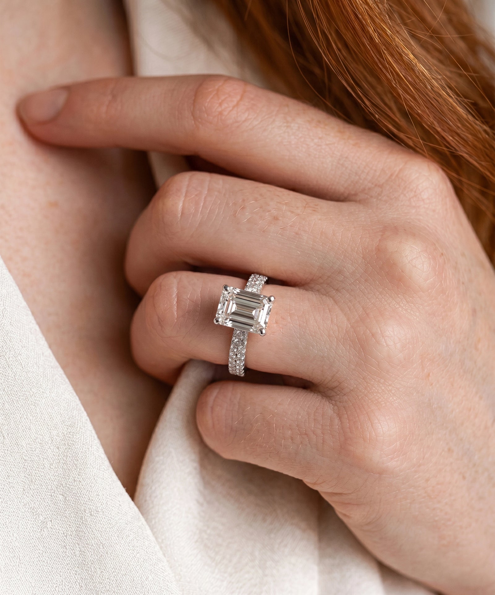 A close-up of a hand wearing a gold ring with an emerald-cut clear center stone and small side stones, set against skin and a soft cream fabric.