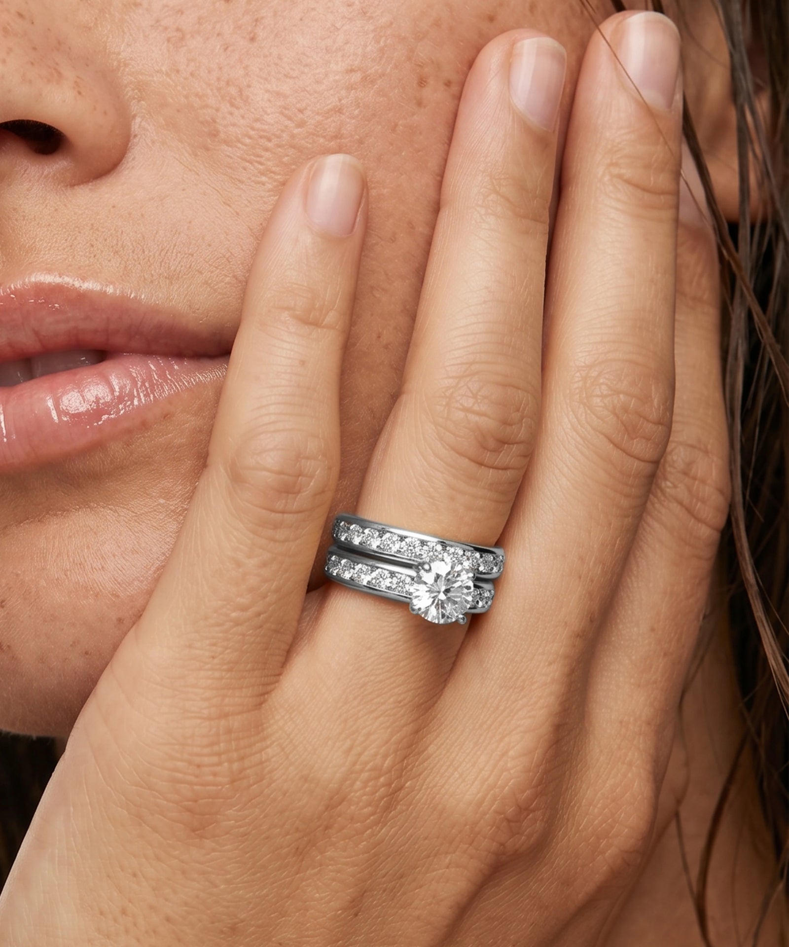 A close-up of a person's hand wearing a silver-toned double-band ring with a round-cut center stone and pavé-set side stones, against a close-up of the model's face.