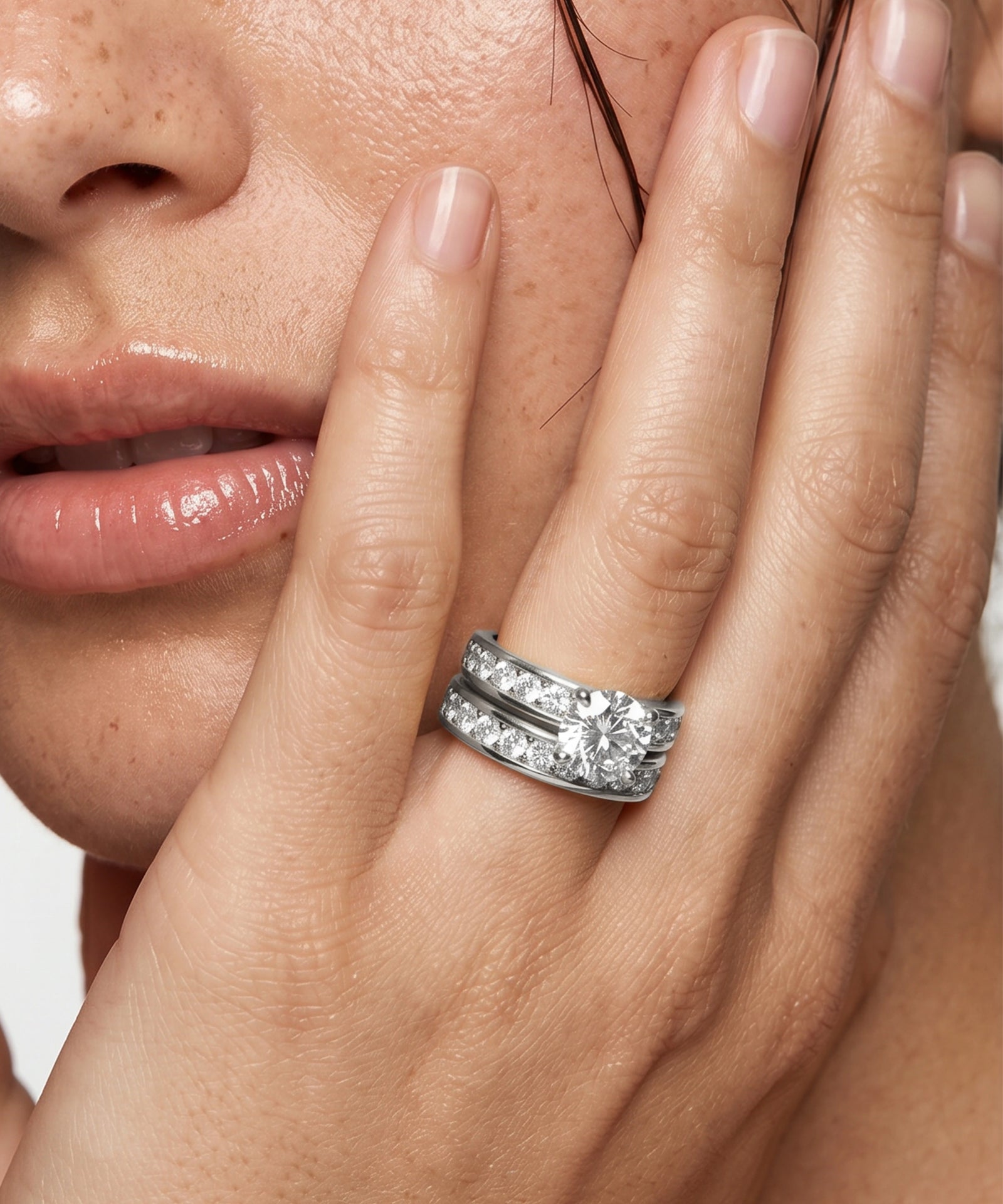A close-up of a woman's hand and lower face wearing a silver double-band ring set with clear round-cut gemstones. The ring is shown on her middle finger against a clean white background.