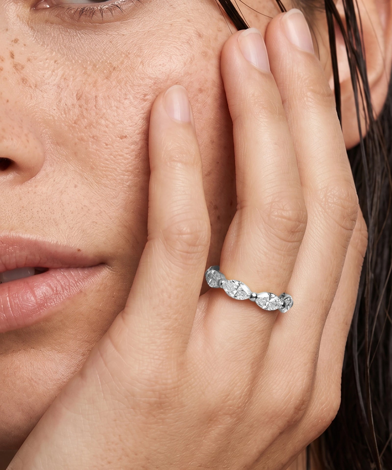 A close-up lifestyle shot of a woman's hand resting against her cheek, wearing a silver ring with a row of marquise-cut clear gemstones.