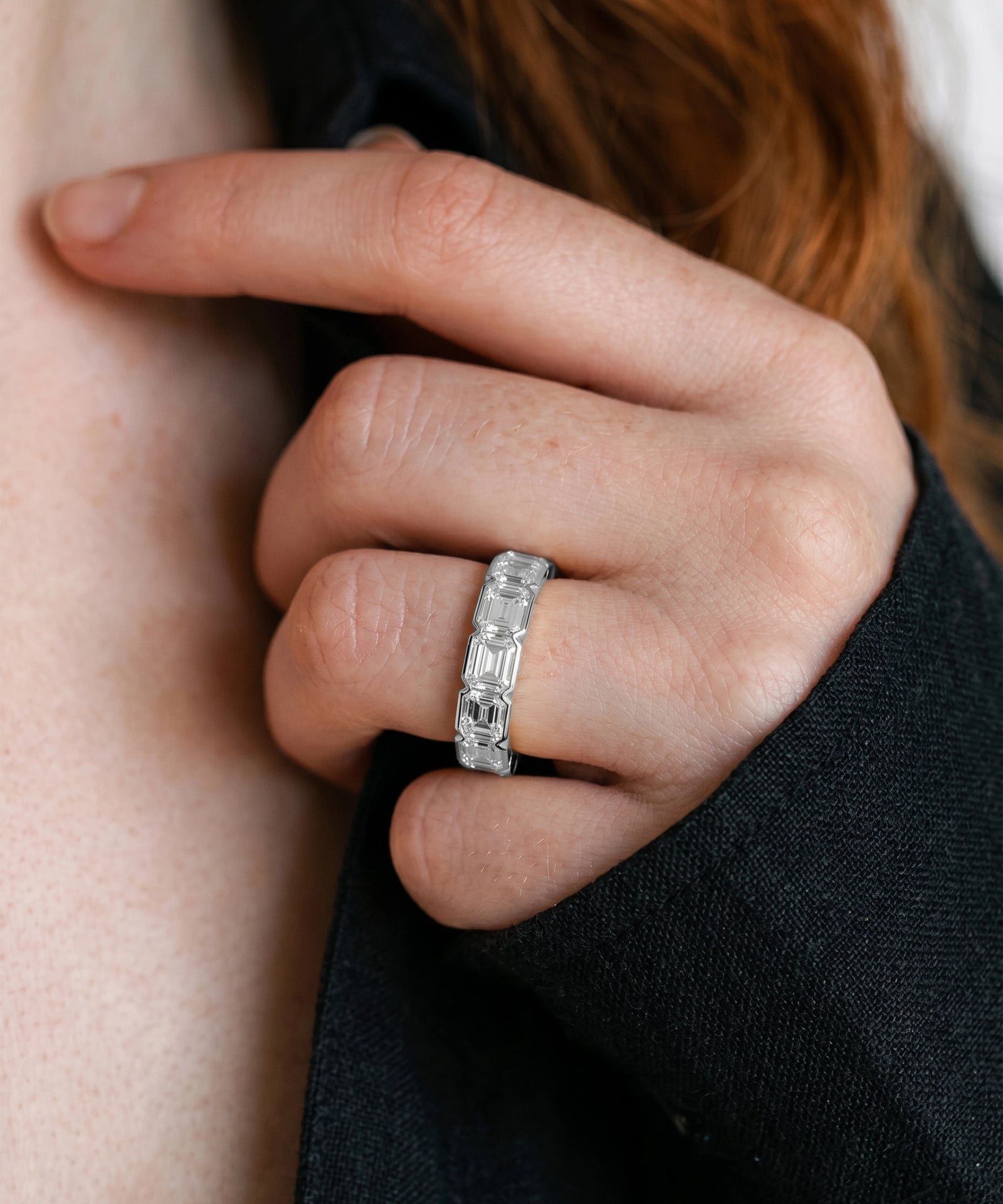 A close-up of a person's hand wearing a silver band ring with baguette-cut clear stones, set against a dark sleeve and skin tone.