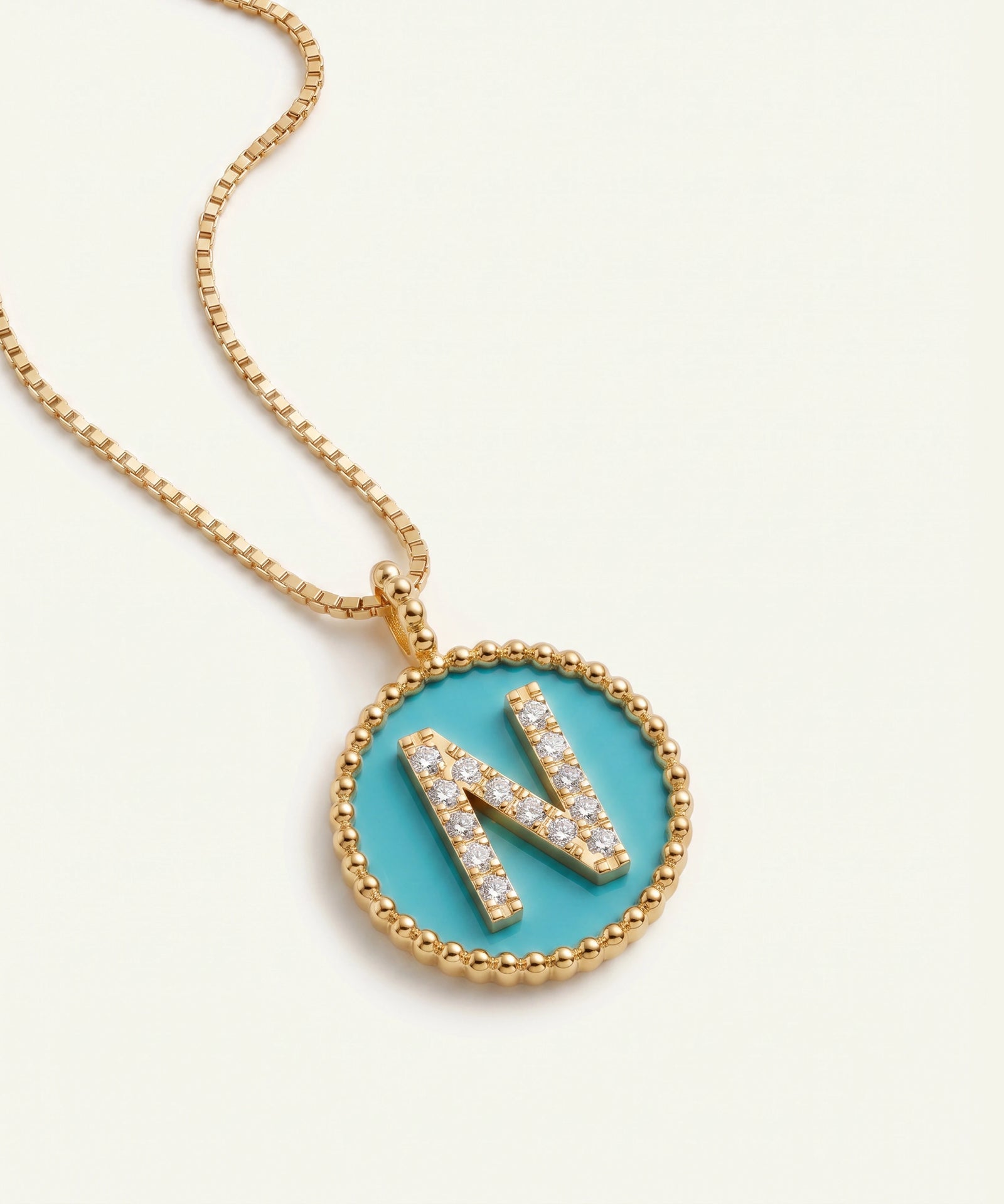 A gold chain necklace with a round bezel pendant featuring a turquoise blue center and a raised letter 'N' paved with small clear stones, shown on a light cream background.