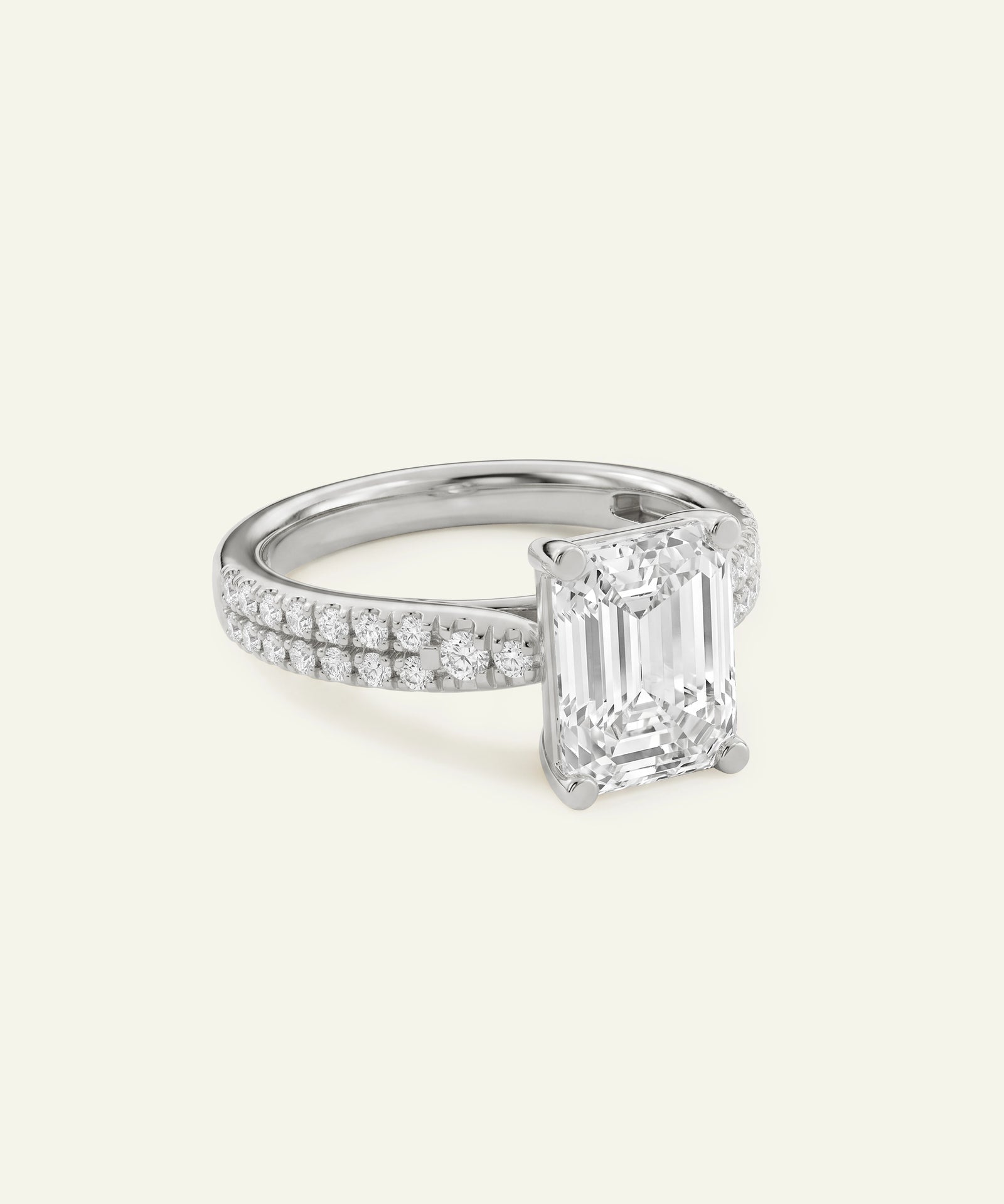 An emerald-cut diamond engagement ring with a pavé band, set in a silver-colored metal on a light cream background.