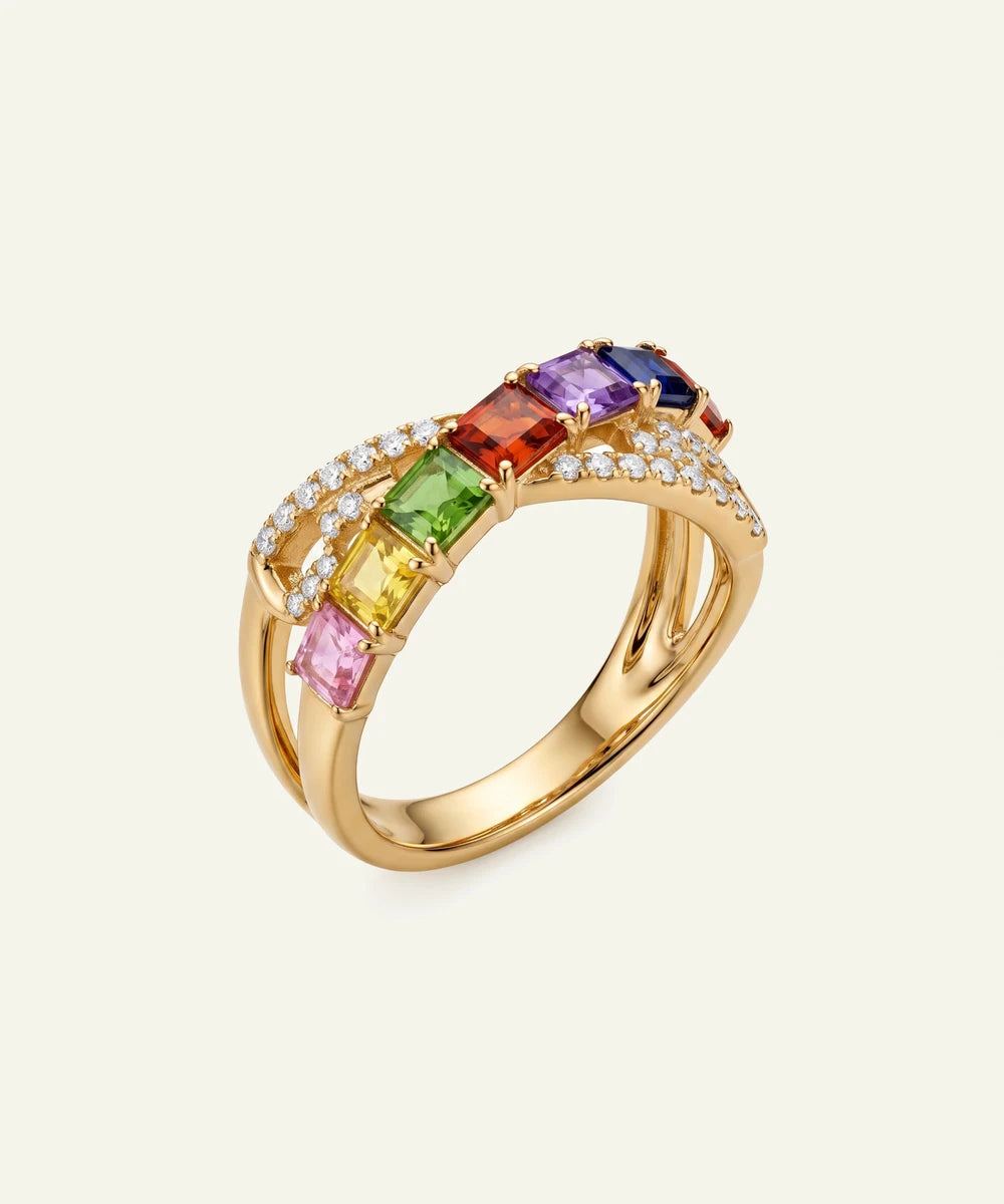 A gold-plated double-band ring featuring a curved top set with rainbow-colored gemstones (pink, yellow, green, orange, purple) and small clear accent stones, shown on a light cream background.
